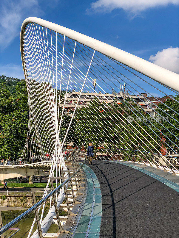 行人在未来主义的Campo Volantin人行桥上穿过Nervion河，桥上有一个系拱设计和弯曲的人行道